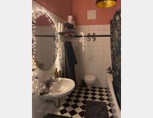 Kleines Badezimmer mit weiß gefliesten Wänden, schwarz-weiß kariertem Boden und einer rosa gestrichenen Decke. Ein ovaler Spiegel mit Lichterkette hängt über einem Waschbecken, daneben befinden sich Handtücher, Regale und eine Toilette; rechts ein Duschvorhang mit Pflanzenmuster.