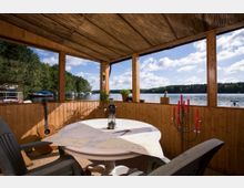 Überdachte Terrasse aus Holz mit Blick auf einen See; im Vordergrund ein runder Tisch mit einer Landkarte, einer Laterne und Stühlen, daneben eine Kerze im roten Kerzenständer. Im Hintergrund sind Bäume, Boote und der blaue Himmel mit weißen Wolken zu sehen.