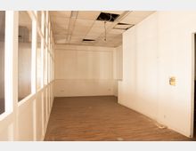 Leerer Innenraum eines ehemaligen Büro- oder Gewerberaums mit weiß gestrichenen Wänden, braunem Laminatboden und einer abgehängten Rasterdecke mit offenen Deckenfeldern und herabhängenden Kabeln. Links verläuft eine Glaswand mit Fenstern, rechts ist eine Öffnung bzw. ein Zugang in der Wand zu sehen.