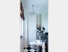 Kleines Badezimmer mit schwarz-weiß kariertem Fliesenboden, weißem Waschbecken, einer Toilette und einem schwarzen Waschtisch. Ein Kronleuchter hängt von der Decke, während Einbauschränke und offene Regale mit dekorativen Elementen, wie Kerzen und Pferdefiguren, den Raum schmücken.