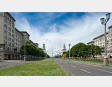 Breite Straße mit begrüntem Mittelstreifen, flankiert von symmetrischen, historischen Wohngebäuden im Stil des sozialistischen Klassizismus. Im Hintergrund sind zwei Türme mit grünen Kuppeln sowie der Fernsehturm zu sehen, der in den bewölkten Himmel ragt.