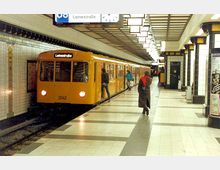 Eine gelbe U-Bahn der Linie U8 steht an einer unterirdischen Station mit der Aufschrift "Leinestraße". Die Wände sind mit weißen Fliesen verkleidet, es gibt schwarze und gelbe Verzierungen an den Säulen, und eine helle Beleuchtung hängt von der Decke. Ein Fahrgast betritt die Bahn, während andere auf dem Bahnsteig gehen.