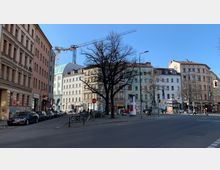 Straßenszene in einer urbanen Umgebung mit mehrstöckigen Altbauten, teils mit pastellfarbenen Fassaden. Ein zentraler Platz mit Bäumen, parkenden Autos, Fahrrädern und einem Baukran im Hintergrund vor klarem, blauem Himmel.