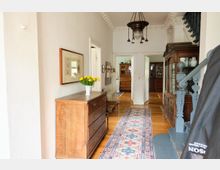 Heller Flur in einem historischen Haus mit Holzboden, weiß gestrichenen Wänden und einer verzierten Deckenleiste. Zu sehen sind ein Holztresen mit einer Vase gelber Blumen, ein persischer Läufer, ein antikes Möbelstück mit Glasvitrine, eine Holztreppe sowie offene Türen, die in andere Räume führen.