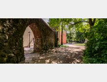 Steinbogen eines historischen Gebäudes, flankiert von einer alten Backsteinmauer, führt zu einem Pfad, der durch eine grüne, baumreiche Parklandschaft verläuft. Im Hintergrund sind ein rotes Gebäude und eine Laterne sichtbar.