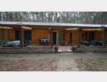 Ein einstöckiges Holzhaus mit überdachter Terrasse, umgeben von einem Wald. Die Terrasse ist mit Sitzbänken, einem Tisch mit Tischdecke, einem Grill und abgestellten Fahrrädern ausgestattet, daneben ein gepflasterter Vorhof.