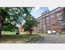 Industriegelände mit einer großen, mehrstöckigen Backsteinfassade, die zahlreiche vergitterte Fenster aufweist, einige davon beschädigt. Im Vordergrund ein gepflasterter Weg, Bäume und Wiesenflächen; im Hintergrund stehen Container und Mülltonnen.