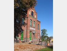 Backsteinbahnhofsgebäude mit grün gestrichenen Fenster- und Türrahmen, einem markanten Uhrturm und einem Schild mit der Aufschrift "Mellen-Saalow". Im Vordergrund sind herbstliche Blätter, Pflanzkübel und ein Dreibein-Stativ sichtbar, umgeben von einer ländlichen Umgebung.