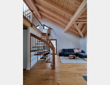 Modernes Wohnzimmer mit offenem Holzgebälk und sichtbarem Dachstuhl. Eine Holztreppe mit Metallgeländer führt zu einer Empore, daneben eine Sitzecke mit dunkelgrauem Sofa, hellem Teppich und Holztisch auf einem Holzboden.