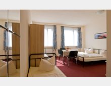 Ein einfacher Mehrbettzimmerraum mit Teppichboden in Rot, zwei Betten mit Metallrahmen, einem schlichten Holztisch und vier schwarzen Stühlen. Vorhänge mit buntem Muster rahmen die Fenster, durch die Tageslicht einfällt; ein kleines Wandbild hängt an der beigefarbenen Wand.