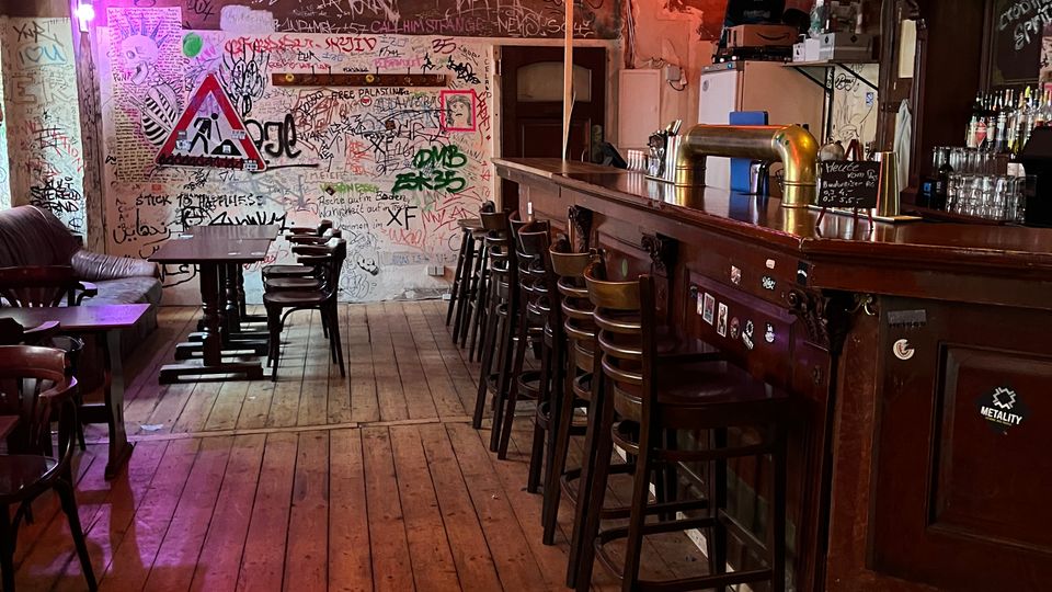 Innenraum einer rustikalen Bar mit Holzdielenboden, langen Tresen mit Barhockern und Regalen mit Flaschen. Die Wände sind dicht mit Graffiti und Stickern bedeckt, dazu hängen Schilder und warme Deckenlampen, teils in farbigem Licht.