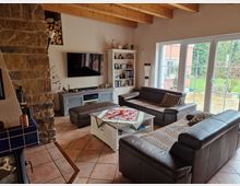 Gemütliches Wohnzimmer mit Ledersofas, Couchtisch und Kamin aus Naturstein. Große Fensterfront mit Blick auf den Garten, Fliesenboden und sichtbare Holzdeckenbalken betonen die warme Atmosphäre des Raumes.