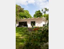 Ein einstöckiges Gebäude mit wellenförmigem Dach und großen Fenstern, umgeben von einer grünen, bewachsenen Landschaft. Im Vordergrund sind eine Rasenfläche, Blumenbeete und eine kleine Treppe zu sehen, während im Hintergrund dichte Bäume stehen.