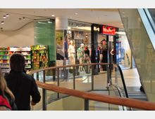 Einkaufszentrum mit einem geschwungenen Geländer aus Holz und Glas neben einer Rolltreppe. Im Hintergrund sind ein Bekleidungsgeschäft und Regale mit Produkten aus einem Lebensmittelgeschäft zu sehen.