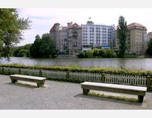 Ein Uferweg mit zwei Holzbänken und einem niedrigen weißen Zaun im Vordergrund; dahinter ein ruhiger See, umgeben von grünen Bäumen und mehrstöckigen Gebäuden, darunter ein Hotel mit der Aufschrift „Seehof“ und einer deutschen Flagge auf dem Dach.