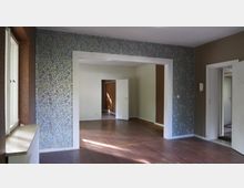 Ein leerer Innenraum eines Hauses mit Holzboden, teilweise tapezierten Wänden in floralem Muster und weißen Wänden. Durch eine offene Tür und Rahmen sind weitere Zimmer zu sehen, eines mit Holzvertäfelung.