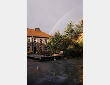 Hinterhof mit einem Ziegelgebäude und rotem Schrägdach, umgeben von Bäumen. Im Vordergrund eine gepflasterte Fläche mit Pfützen, im Hintergrund ein Regenbogen am bewölkten Himmel.