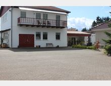 Zweistöckiges Wohnhaus mit weißer Fassade, rotem Satteldach und einem Balkon mit dekorativem Geländer. Im Vordergrund befindet sich eine gepflasterte Auffahrt, daneben kleinere Pflanzen und ein angrenzender Anbau mit überdachtem Eingang.