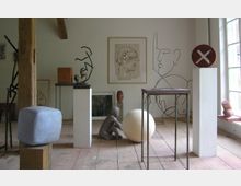 Ein Atelierraum mit Holzboden, weißen Wänden und zahlreichen Kunstobjekten, darunter Skulpturen aus Metall, Stein und Ton sowie Zeichnungen an der Wand. Hölzerne und metallene Sockel sowie eine große Fensterfront sorgen für eine helle, kreative Atmosphäre.