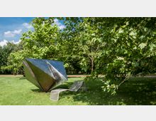 Eine moderne Metallskulptur in geometrischer Form steht auf einer Rasenfläche in einem Park, daneben eine geschwungene Metallbank. Umgeben von grünen Bäumen, die im Sonnenlicht leuchten, erstreckt sich der Park bis zu einem Weg im Hintergrund.