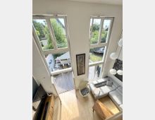 Heller, zweigeschossiger Wohnraum mit hohen Decken, bodentiefen Fenstern und einer Glastür, die auf eine Holzterrasse und in den Garten führt. Im Raum stehen ein graues Ecksofa, ein Couchtisch aus Holz, ein TV-Board und eine hängende Kugelleuchte.