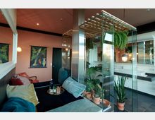 Modern eingerichteter Raum mit einer Sitzecke aus Polstern und Kissen, umgeben von Glaswänden und zahlreichen Topfpflanzen. Im Hintergrund sind eine rosa Wand mit botanischen Wandbildern und ein Waschbecken mit Spiegel zu sehen.