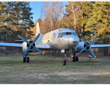 Silbernes Propellerflugzeug mit vier Propellern, geparkt auf einer Wiese vor einem Hintergrund aus hohen Nadel- und Laubbäumen bei Tageslicht.