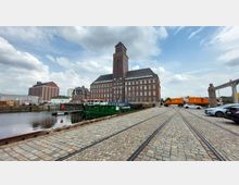 Großes Backsteingebäude mit Turm in einem Hafengebiet, umgeben von gepflasterten Straßen und Schienen. Im Vordergrund ein Kanal mit einem grünen Schiff, im Hintergrund geparkte Fahrzeuge und industrielle Anlagen.