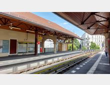 Historischer S-Bahnhof mit einem Bahnsteig, überdacht durch ein hölzernes Dach mit metallischen Stützen. Im Hintergrund sind ein Zugangsbogen, Infotafeln, eine Uhr und benachbarte Gebäude zu sehen, daneben verlaufen Gleise und Signalmarkierungen.