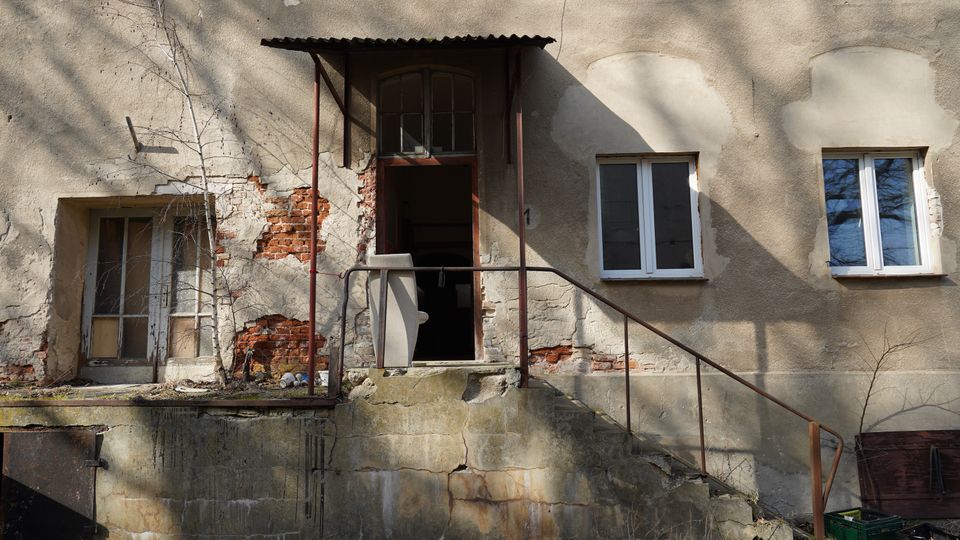 Eingangsbereich eines stark verwitterten Gebäudes mit bröckelndem Putz und freiliegendem Ziegelmauerwerk, einer offenen Tür unter einem kleinen Wellblechvordach. Eine betonierte Rampe mit rostigem Geländer führt zur Tür; daneben sind alte Fenster und überwucherte Bereiche am Sockel zu sehen.