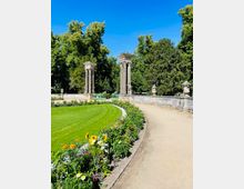 Parkanlage mit geschwungenem Kiesweg entlang eines Blumenbeets und einer großen Rasenfläche. Im Hintergrund stehen zwei hohe steinerne Säulenmonumente neben einer niedrigen Balustrade, umgeben von dichtem Baumbestand unter blauem Himmel.