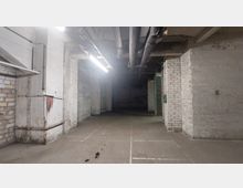 Ein leerer, unterirdischer Raum mit weißen Ziegelwänden, Betonboden und sichtbaren Rohren an der Decke. Schwaches Licht fällt von einer langen Leuchtstoffröhre, und der hintere Bereich ist dunkel.