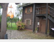 Holzgebäude mit Blockhausoptik, das als Museum dient, mit einem Eingang im Erdgeschoss, einer Außentreppe zu einer oberen Tür und Holzfenstern mit Fensterläden. Im Vordergrund ein gepflasterter Hof, ein Blumenstrauch in einem Topf und eine grüne Wiese mit Bäumen im Hintergrund.