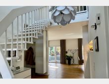 Heller, moderner Wohnungsflur mit offener weißer Treppe und Geländer, Holzfußboden und einer großen, blütenförmigen Pendelleuchte im Luftraum. Im Hintergrund führt eine breite Öffnung in einen Wohnbereich mit großen Fenstern und Vorhängen, Pflanzen und einem schlanken Kerzenständer.