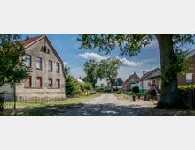 Eine ruhige Dorfstraße mit kleinen Einfamilienhäusern und einem alten, verputzten Gebäude auf der linken Seite. Große Bäume säumen die Straße, und im Hintergrund sind weitere Häuser und blauer Himmel mit Wolken zu sehen.