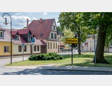 Eine Straßenszene in einem ländlichen Ort mit Fachwerkhäusern und roten Ziegeldächern, umgeben von Bäumen. Im Vordergrund steht ein Schild mit der Aufschrift „Rohdeplatz“ auf einer Grünfläche, daneben befinden sich Parkschilder und eine gepflasterte Straße.