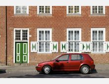Fassade eines Backsteinhauses mit weißen Fenstern und grün-weißen Klappläden, daneben eine Tür in grün-weißem Design; im Vordergrund steht ein rotes Auto am Straßenrand.