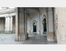 Steinernes, historisches Bauwerk mit Rundbögen und verzierten Reliefs an den Wänden, flankiert von massiven Säulen. Im Hintergrund sind dichte Sträucher und Bäume sichtbar.