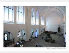 Innenraum eines historischen Gebäudes im Märkischen Museum, Berlin, mit hohen weißen Gewölbedecken, großen gotischen Fensterbögen und steinernem Boden. Zu sehen sind mehrere Ausstellungsstücke, darunter eine große Glocke, Skulpturen und dekorative Elemente.