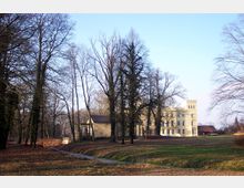 Ein Schloss in klassizistischer Architektur mit heller Fassade und einem auffälligen Turm, umgeben von Bäumen ohne Laub. Im Vordergrund befindet sich ein kleiner Bachlauf mit einer hölzernen Brücke und einer gepflegten Wiesenlandschaft.