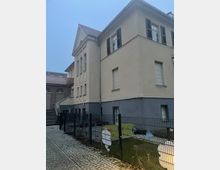 Alt-Text: Außenansicht eines zweistöckigen Gebäudes mit gelblicher Fassade und schwarzen Fensterläden. Im Vordergrund befindet sich ein eingezäunter Bereich mit einer Wiese und Spielplatzdekorationen, während rechts ein gepflasterter Weg verläuft.