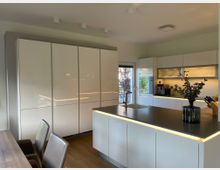 Moderne Küche mit weißen Hochglanzfronten, einer Kochinsel mit dunkler Arbeitsplatte und integriertem Spülbecken. Im Hintergrund befinden sich hohe Schränke, eine Kaffeemaschine, und eine Tür, die zu einem Gartenbereich führt; die Beleuchtung ist dezent in die Möbel integriert.