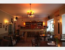 Gemütlicher Café- oder Barraum mit einer Theke im Hintergrund, die mit Flaschen und Gläsern bestückt ist. Der Raum ist mit Holztischen, Stühlen, Sesseln und einer Kronleuchterlampe ausgestattet; an den Wänden befinden sich Bilder und Lampen, während ein Fenster Tageslicht hereinlässt.