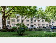 Wohngebäude mit hell verputzter Fassade und mehreren Fenstern, umgeben von Bäumen und einem kleinen Grünstreifen im Vordergrund. Am Straßenrand parken Autos, und es sind Fußgänger sowie ein Straßenschild sichtbar.