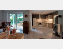 Wohnzimmer mit offener Küche und Zugang zur Terrasse. Im Vordergrund stehen ein Sessel und Dekorationsgegenstände, während die Küche rustikale Holzmöbel, einen weißen Kühlschrank und einen Herd zeigt; eine große Glastür führt auf eine Terrasse mit Garten.