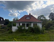 Einfamilienhaus mit weißer Fassade und rotem Ziegeldach mit Schornstein, davor eine Veranda mit großen Fenstern. Umgeben von hohem Gras, dichtem Bewuchs und Bäumen unter einem wolkigen Himmel.