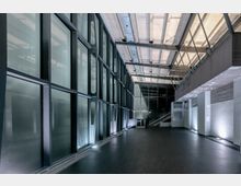 Moderner, minimalistisch gestalteter Innenraum mit großen Glaswänden auf der linken Seite, einer hohen Decke mit sichtbaren Metallträgern und einer Treppe im Hintergrund. Der Boden ist dunkel, und dezente Beleuchtung akzentuiert die Wände und Strukturen.