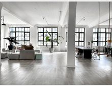 Heller, moderner Wohn- und Arbeitsraum in einem Loft mit großen, schwarz gerahmten Fenstern, weißen Wänden und Holzfußboden. Im Vordergrund befinden sich Sofas und Pflanzen, im Hintergrund ein langer Esstisch mit Stühlen.