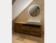 Modernes Badezimmer mit schräger Dachwand, beigefliesten Wänden und Boden sowie einem langen Holzwaschtisch mit Schubladen und rechteckigem Aufsatzwaschbecken. Darüber hängt ein runder, beleuchteter Spiegel; rechts ist eine Steckdose an der Wand zu sehen.