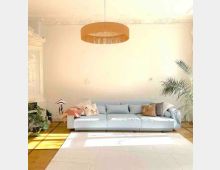 Wohnzimmer mit hellem, großzügigem Interieur: Eine hellblaue, gepolsterte Couch mit dekorativen Kissen steht vor einer weißen Wand mit Stuckverzierungen. Links stehen Pflanzen und eine Tischlampe, rechts eine hohe Palme, der Boden ist mit Parkett und einem hellen Teppich gestaltet, und ein großer, runder Deckenleuchter dominiert den Raum.